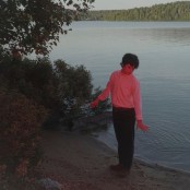 A Man wearing a white long sleeve top, black trousers, brown belt, and brown shows, with curly dark brown hair stands at the edge of a lake on the sand with his back to the water. There is a red hued light shining towards him
